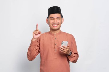 Gülümseyen Asyalı genç Müslüman adam cep telefonunu tutuyor ve işaret ediyor, beyaz arka planda izole edilmiş dahiyane çözümler yaratıyor.