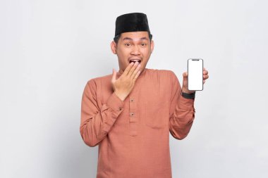 Şaşırmış Asyalı genç Müslüman adam cep telefonu boş ekranı gösteriyor. Beyaz arkaplanda izole edilmiş uygulamayı tavsiye ediyor.