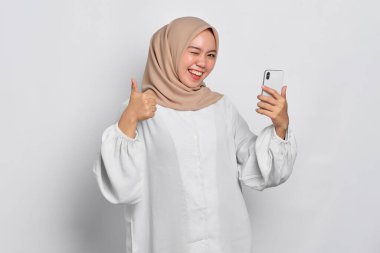 Gülümseyen Asyalı Müslüman kadın cep telefonu tutuyor ve beyaz arka planda izole edilmiş baş parmak hareketi yapıyor.