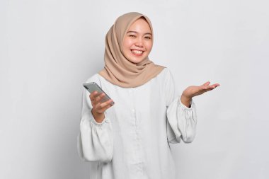 Neşeli Asyalı genç Müslüman kadın cep telefonu kullanıyor ve başarıyı kutluyor, beyaz arka planda iyi haberler alıyor.