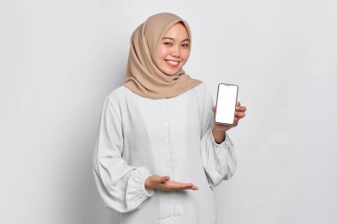Gülümseyen Asyalı Müslüman kadın cep telefonu boş ekranı gösteriyor. Beyaz arka planda izole edilmiş uygulamayı tavsiye ediyor.