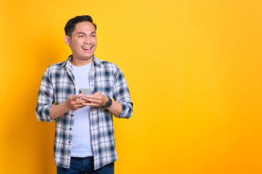 Cheerful young Asian man in plaid shirt holding mobile phone and looking aside at copy space isolated on yellow background