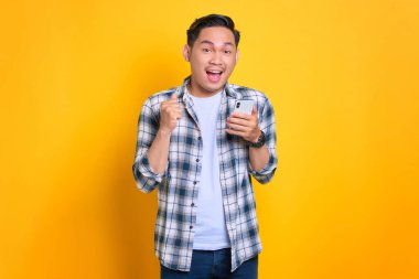 Ekose gömlekli heyecanlı genç Asyalı adam başarıyı cep telefonuyla kutluyor, elini kaldırıyor sarı arka planda izole edilmiş büyük bir şansa seviniyordu.