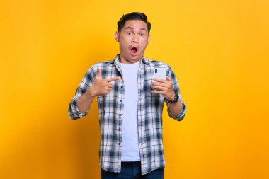 Ekose gömlekli, şok olmuş genç Asyalı bir adam cep telefonunu işaret ederek sarı arka planda izole edilmiş inanılmaz haberler alıyor.