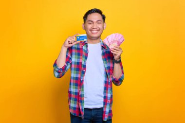 Ekose gömlekli neşeli genç Asyalı adam sarı arka planda para banknotları ve kredi kartı gösteriyor.