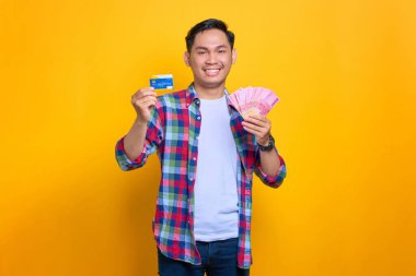 Ekose gömlekli neşeli genç Asyalı adam sarı arka planda para banknotları ve kredi kartı gösteriyor.