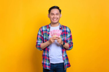 Ekose gömlekli gülümseyen genç Asyalı adam elinde para banknotları ve sarı arka planda izole edilmiş kameraya bakıyor.