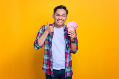 Ekose gömlekli gülümseyen genç Asyalı adam elinde para banknotları ve sarı arka planda izole edilmiş baş parmakları gösteriyor.