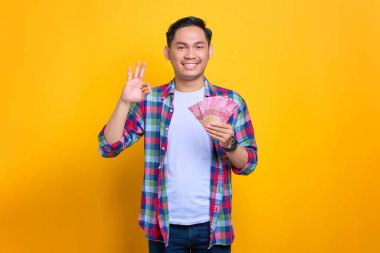 Ekose gömlekli gülümseyen genç Asyalı adam elinde para banknotları ve sarı arka planda izole edilmiş işaret işareti gösteriyor.