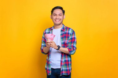 Ekose gömlekli gülümseyen genç Asyalı adam elinde para banknotları ve sarı arka planda izole edilmiş kameraya bakıyor.