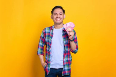 Ekose gömlekli gülümseyen genç Asyalı adam elinde para banknotları ve sarı arka planda izole edilmiş kameraya bakıyor.