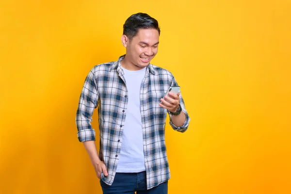 Ekose gömlekli gülümseyen genç Asyalı cep telefonu kullanıyor. Sarı arka planda izole edilmiş iyi habere tepki veriyor.