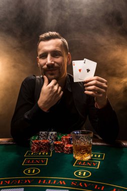 Elinde iki as olan yakışıklı bir poker oyuncusu sigara dumanıyla dolu karanlık bir odada poker masasında oturuyor..
