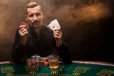 Elinde iki as olan yakışıklı bir poker oyuncusu sigara dumanıyla dolu karanlık bir odada poker masasında oturuyor..
