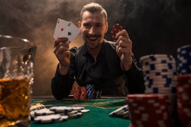 Elinde iki as olan yakışıklı bir poker oyuncusu sigara dumanıyla dolu karanlık bir odada poker masasında oturuyor..