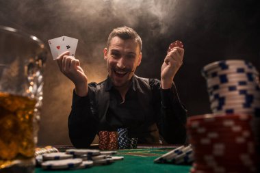 Elinde iki as olan yakışıklı bir poker oyuncusu sigara dumanıyla dolu karanlık bir odada poker masasında oturuyor..