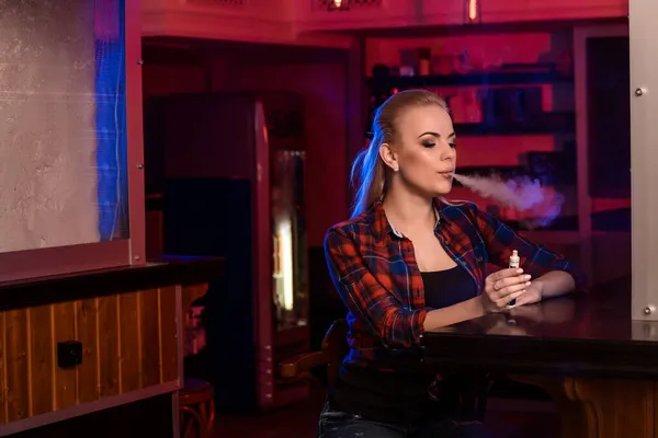 Joven mujer bonita en una camisa en una jaula fumar un cigarrillo electrónico en la barra de vapor — Foto de Stock