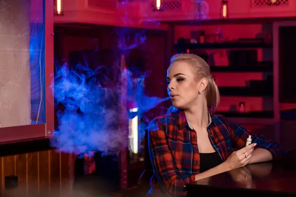 Νεαρή όμορφη γυναίκα σε ένα πουκάμισο σε ένα κλουβί καπνίζει ένα ηλεκτρονικό τσιγάρο στο μπαρ — Φωτογραφία Αρχείου