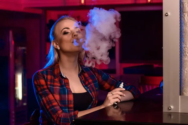 Νεαρή όμορφη γυναίκα σε ένα πουκάμισο σε ένα κλουβί καπνίζει ένα ηλεκτρονικό τσιγάρο στο μπαρ — Φωτογραφία Αρχείου