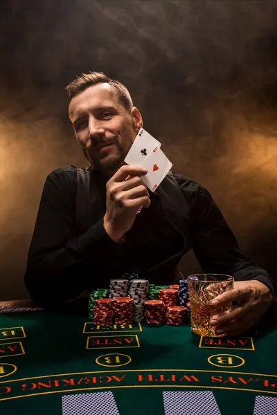 Guapo jugador de póquer con dos ases en sus manos y fichas sentado en la mesa de póquer en un cuarto oscuro lleno de humo de cigarrillo. — Foto de Stock