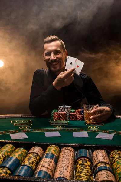 Bello giocatore di poker con due assi nelle mani e patatine fritte seduto al tavolo da poker in una stanza buia piena di fumo di sigaretta. — Foto Stock