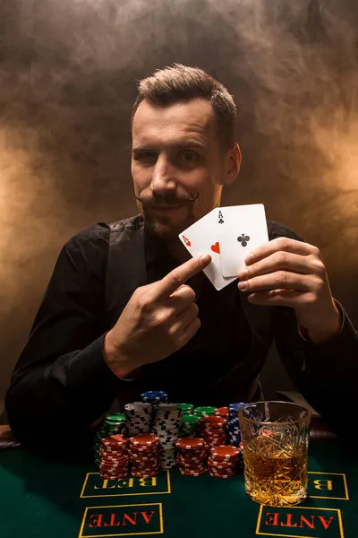 Guapo jugador de póquer con dos ases en sus manos y fichas sentado en la mesa de póquer en un cuarto oscuro lleno de humo de cigarrillo. — Foto de Stock