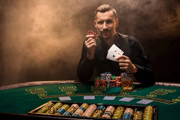 Knappe pokerspeler met twee azen in zijn handen en chips zittend aan pokertafel in een donkere kamer vol sigarettenrook. — Stockfoto