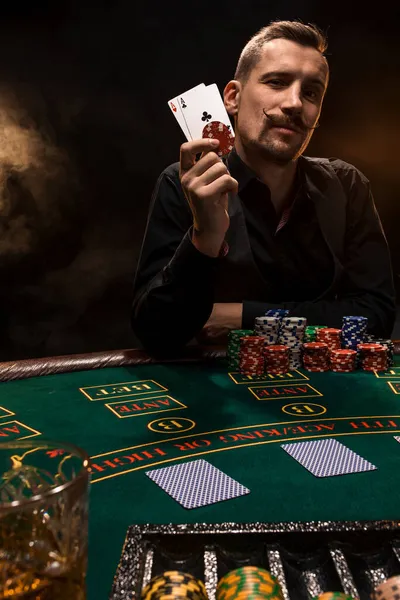 Bello giocatore di poker con due assi nelle mani e patatine fritte seduto al tavolo da poker in una stanza buia piena di fumo di sigaretta. — Foto Stock