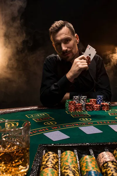 Beau joueur de poker avec deux as dans les mains et des jetons assis à la table de poker dans une pièce sombre pleine de fumée de cigarette. — Photo