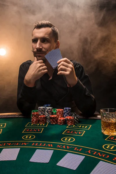 Schöner Pokerspieler mit zwei Assen in den Händen und Chips am Pokertisch in einem dunklen Raum voller Zigarettenrauch. — Stockfoto