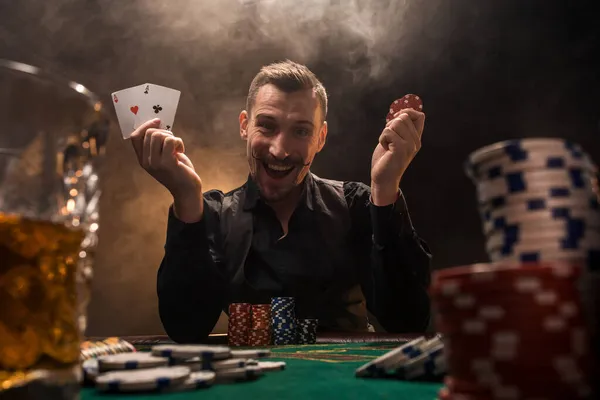 Guapo jugador de póquer con dos ases en sus manos y fichas sentado en la mesa de póquer en un cuarto oscuro lleno de humo de cigarrillo. —  Fotos de Stock