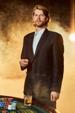 Oyun masasının yanında takım elbiseli bir adam duruyor. Erkek oyuncu. Tutku, kartlar, fişler, alkol, zar, kumar, kumarhane - erkek eğlencesi gibi.