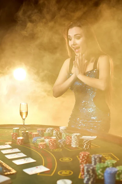 Mujer hermosa joven posa cerca de la mesa de póquer en el casino de lujo. Pasión, cartas, fichas, alcohol, dados, juegos de azar, casino es como entretenimiento femenino. Fondo de humo. — Foto de Stock