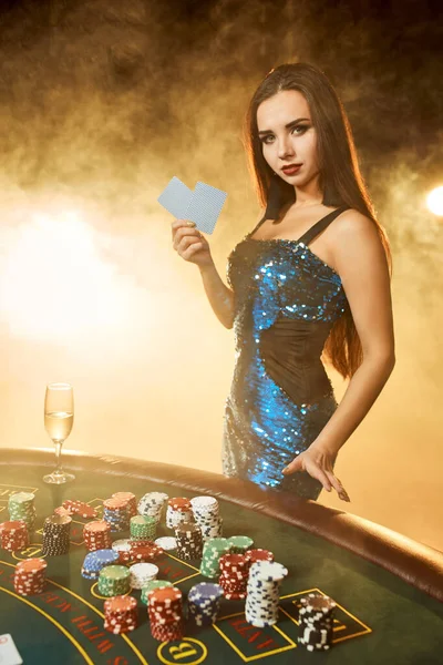 Genç ve güzel bir kadın lüks kumarhanede poker masasının yanında poz veriyor. Tutku, kartlar, fişler, alkol, zar, kumar, kumarhane, hepsi kadın eğlencesi. Duman arkaplanı. — Stok fotoğraf