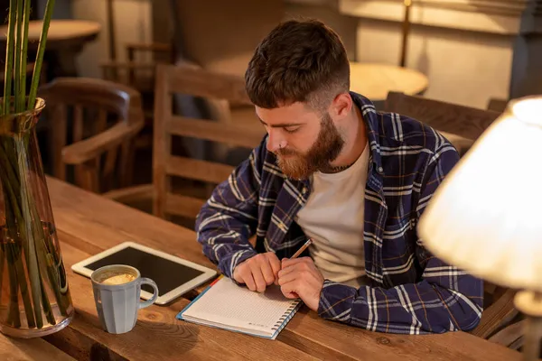 Der junge bärtige Geschäftsmann sitzt im Café, zu Hause am Tisch und schreibt in Notizbücher, daneben liegt ein Tablet-Computer mit schwarzem Bildschirm. Mann arbeitet, studiert. — Stockfoto