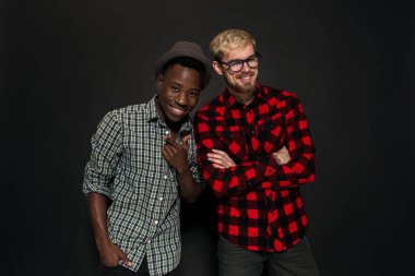 Stüdyo çekimlerinde iki şık genç adam eğleniyor. Kafesteki tişörtlü yakışıklı sakallı hippi, siyah arka planda şapkalı Afro-Amerikan arkadaşının yanında duruyor..