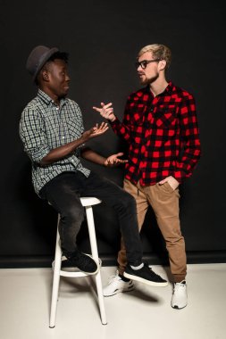 Stüdyo çekimlerinde iki şık genç adam eğleniyor. Kafesteki tişörtlü yakışıklı sakallı hippi, siyah arka planda şapkalı Afro-Amerikan arkadaşının yanında duruyor..