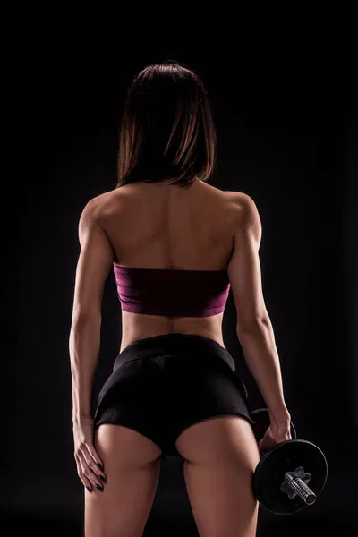 Сильная мускулистая молодая женщина держит гантели на боку, стоя спиной к камере, глядя в сторону, на черном с копировальным пространством — стоковое фото