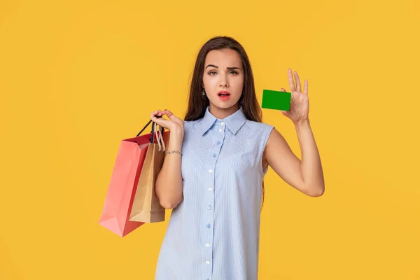 Mujer de moda de 20 años en vestido con el pelo largo marrón sonriendo mientras sostiene diferentes paquetes de compras y tarjeta en las manos aisladas sobre fondo amarillo — Foto de Stock