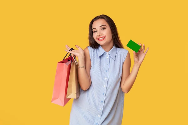 Mujer de moda de 20 años en vestido con el pelo largo marrón sonriendo mientras sostiene diferentes paquetes de compras y tarjeta en las manos aisladas sobre fondo amarillo — Foto de Stock