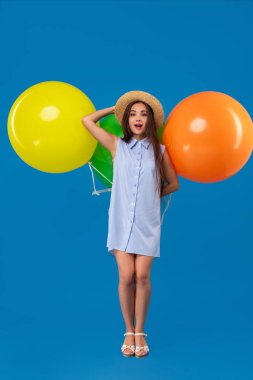 Şirin esmer ayakta turuncu, sarı ve yeşil balonlar tutan bir stüdyoda. O ışık mavi elbise giyer ve uzun ve dalgalı saçları var..