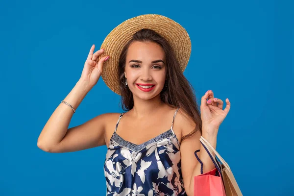 Elinde alışveriş çantaları olan ve mavi arka planda izole edilmiş kameraya bakan yaz şapkalı bir kadının portresi — Stok fotoğraf