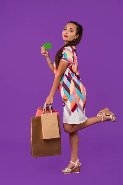 Привлекательная женщина 20 лет в платье с пакетами покупок и показывая зеленую кредитную карту изолированы на фиолетовом фоне — стоковое фото