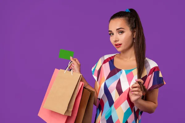 Mujer atractiva 20s en vestido sosteniendo paquetes de compras y mostrando tarjeta de crédito verde aislado sobre fondo púrpura — Foto de Stock