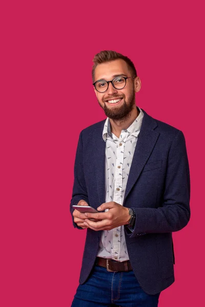 Νεαρός χαρούμενος δικηγόρος ή επιχειρηματίας στέκεται στο ροζ φόντο και χαμογελώντας, διαβάζοντας καλά νέα στο διαδίκτυο στο κινητό του τηλέφωνο. — Φωτογραφία Αρχείου