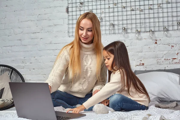 Красива молода мати і її мила дочка в білих светрах і джинсах лежать на ліжку вдома, сміючись і дивлячись в ноутбук — стокове фото