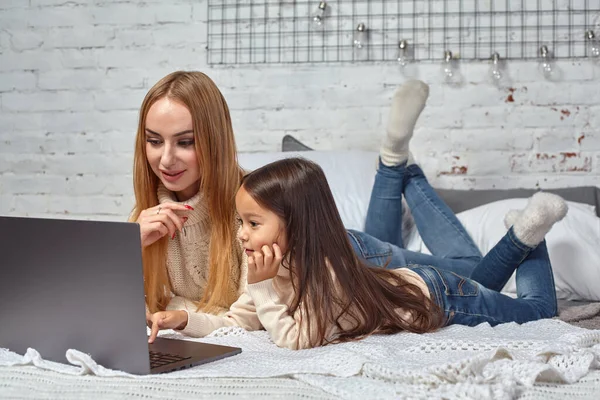 Красивая молодая мать и ее милая дочь в белых свитерах и джинсах лежат на кровати дома, смеясь и глядя в ноутбук — стоковое фото