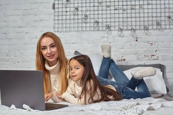 Красивая молодая мать и ее милая дочь в белых свитерах и джинсах лежат на кровати дома, смеясь и глядя в ноутбук — стоковое фото