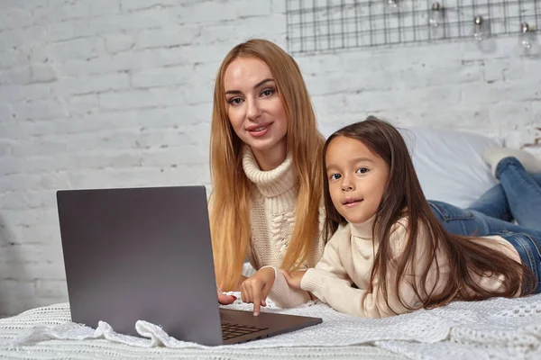Красива молода мати і її мила дочка в білих светрах і джинсах лежать на ліжку вдома, сміючись і дивлячись в ноутбук — стокове фото