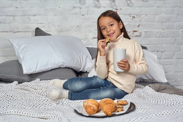 Niña bonita de 6 años con un suéter blanco y jeans. Niño en la habitación con una cama, comer croissant y beber té o cacao. —  Fotos de Stock